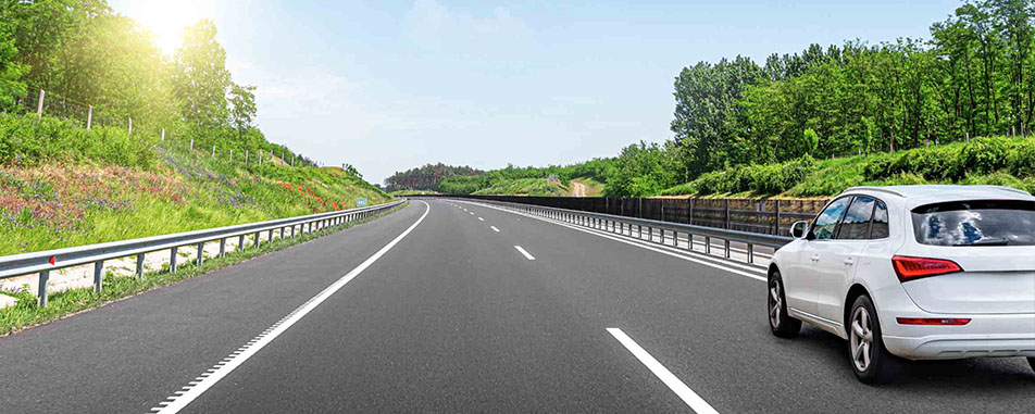 A white car traveling along a highway, surrounded by green trees and blue sky.